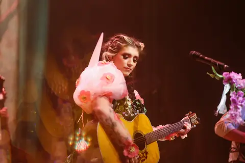 A musician wearing a floral dress plays an acoustic guitar on stage.