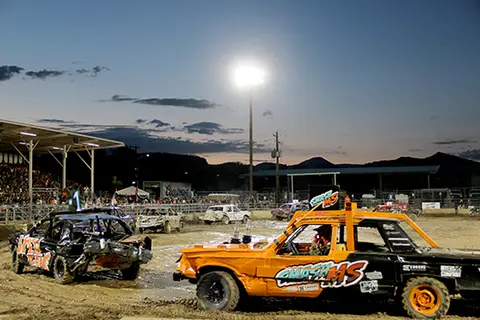 A demolition derby event taking place at dusk with wrecked cars on a dirt track and a crowd watching from a grandstand.