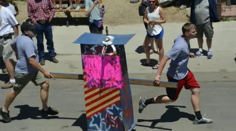 Two people carry a decorated box structure outside on a street.