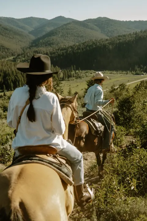 Two people ride horses on a trail through a mountain landscape.