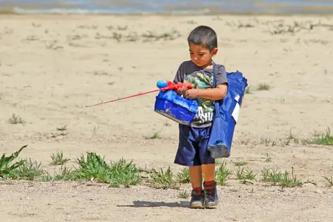 A young boy carries a fishing rod and gear while walking on sandy ground.