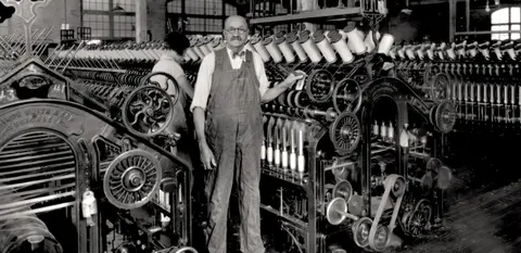 A historic black and white photograph showing a man operating a large textile machine in a factory.