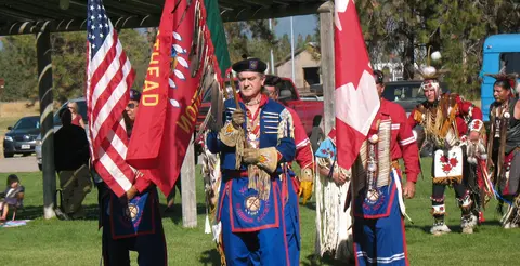 People carrying flags and wearing traditional regalia outdoors.