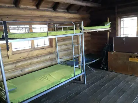 A rustic log cabin interior features metal bunk beds with green mattresses and wooden walls.