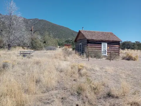 A rustic log cabin sits in a dry grassy field with mountains in the distance.