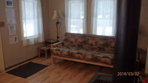 An interior view of a room with a patterned futon sofa, windows with white curtains, and a wooden floor.