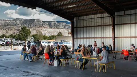 People gather at tables under a large open-sided metal structure with mountains visible in the distance.
