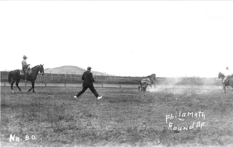 Black and white historic photo of a roundup event with horses and people in an outdoor field.
