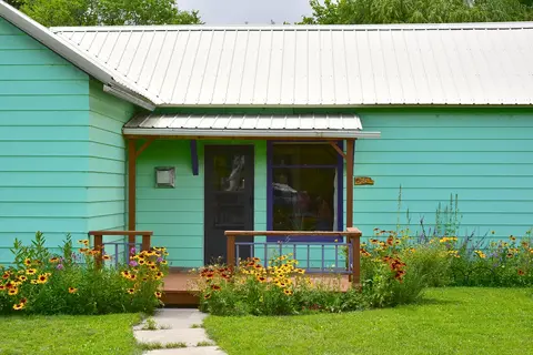 A brightly painted green house with a white metal roof and a small front porch surrounded by flowers.