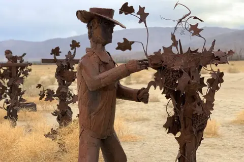 A metal sculpture of a farmer harvesting grapes stands in a dry landscape.