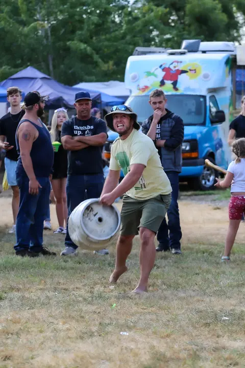 A man carries a metal keg outdoors while spectators watch.