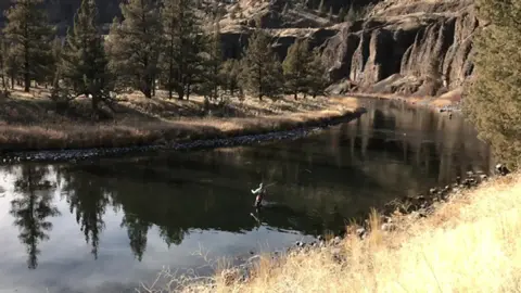 A person wades and fishes in a river bordered by rocky cliffs and sparse trees.