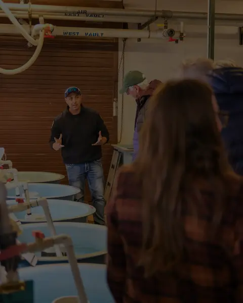 A man speaks to a group of people inside an industrial facility with round tanks.