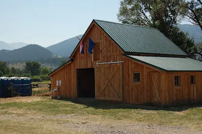 A wooden barn sits in a rural field with mountains in the background.
