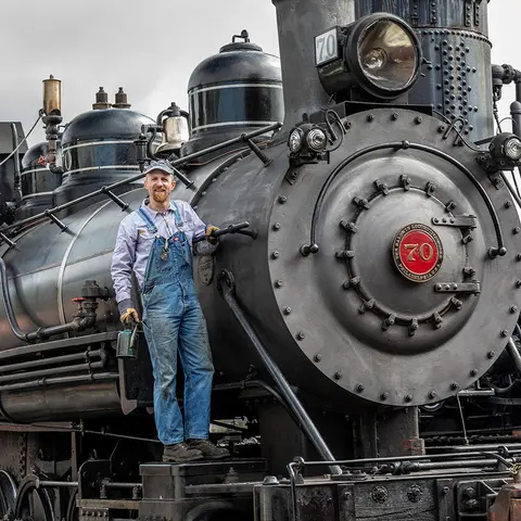 A man in overalls stands on a steam locomotive.