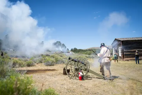 A person in historical attire fires a cannon outdoors with smoke billowing.