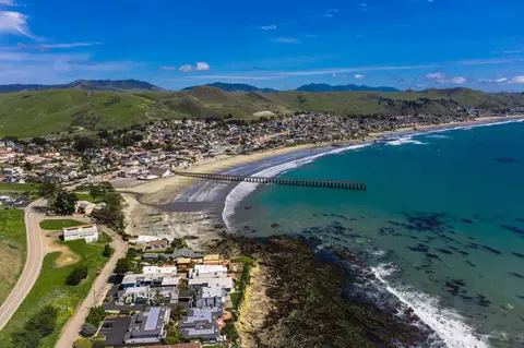 An aerial view shows a coastal town, beach, pier, and surrounding hills.