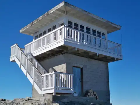 A concrete fire lookout tower with white railings and an outdoor staircase against a blue sky.