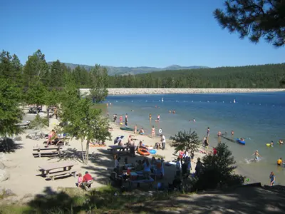 People enjoy a sunny day at a sandy beach beside a lake with a dam in the background.