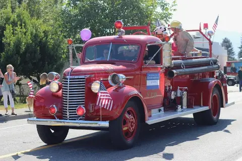 An old red fire truck drives down a street during a parade with flags attached.
