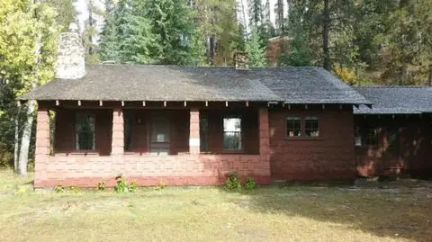 A rustic single-story cabin with a porch and stone chimneys nestled among trees.