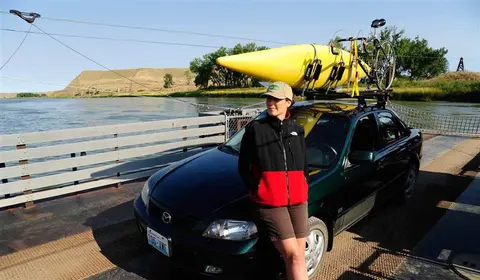 A woman leaning against a car loaded with a kayak and a bicycle on a ferry.