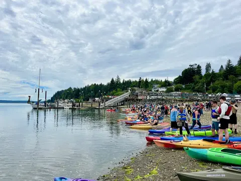 Colorful kayaks are lined up on a shoreline alongside people gathered by the water.