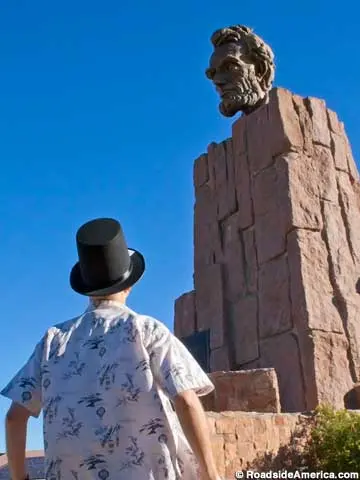 A person wearing a top hat looks up at a large bust sculpture of a historical figure.