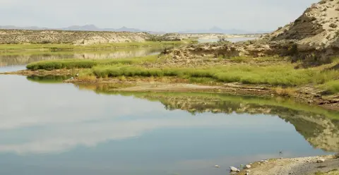 A calm body of water reflects arid landscape and distant mountains.