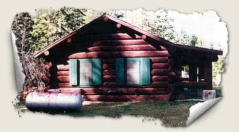 A log cabin with green shutters stands among trees.