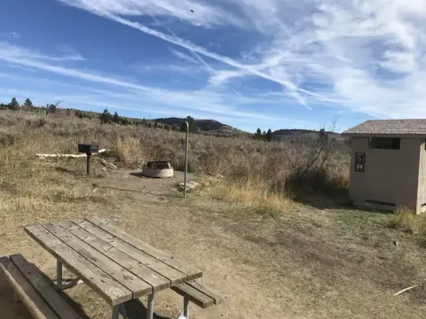 A picnic area with a wooden table, fire ring, grill, and restroom facility under a cloudy sky.