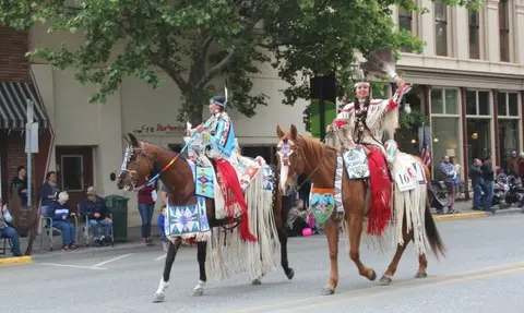 Two women in traditional attire ride horses in a parade.