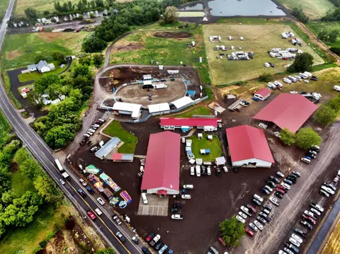 An aerial view of buildings, parking lots, and a dirt arena in a rural area.
