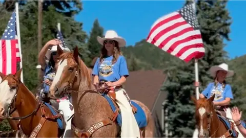 People on horseback carry American flags outdoors.