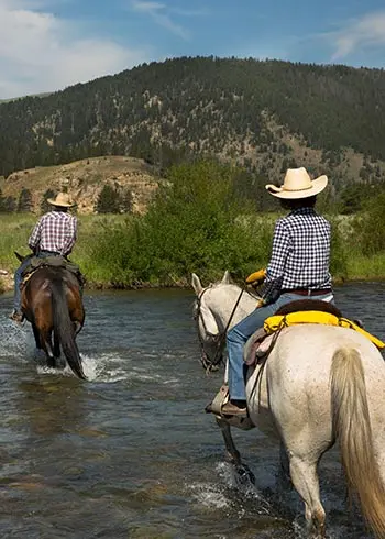 Two people ride horses across a river with a mountain in the background.