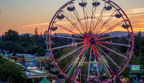 A brightly lit Ferris wheel at an evening fair with mountains in the background.
