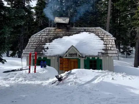 A snowy wooden cabin with smoke coming from the chimney and skis propped outside.