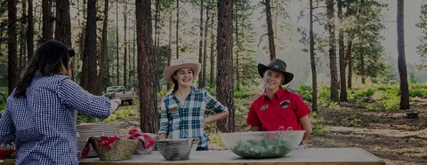 Two women wearing cowboy hats stand behind a buffet table outdoors in a forest.