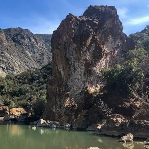 A large rock formation stands by a body of water in a mountainous landscape.