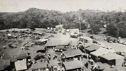 An elevated historic photograph shows an outdoor market or fair with tents, cars, and visitors.