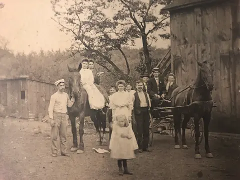 Historic photograph of people posing with horses and a carriage near a barn.