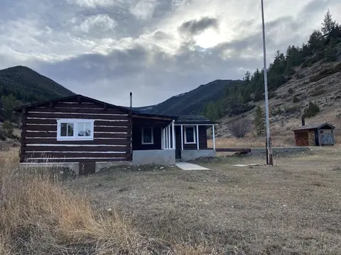 A dark wooden cabin with a covered porch stands in a grassy valley beneath rolling hills.