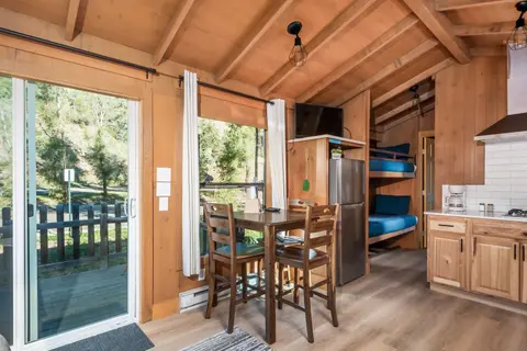 An interior view of a wooden cabin featuring a kitchen, dining table, and bunk beds.
