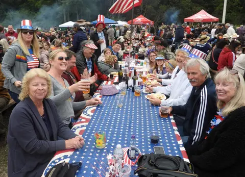 A large crowd of people gathered at outdoor tables with patriotic tablecloths.