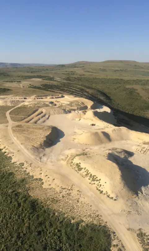 An aerial view of an open-pit quarry surrounded by green landscape under a clear sky.