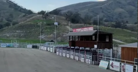 An outdoor rodeo arena with a wooden judge's booth and rolling green hills in the background.