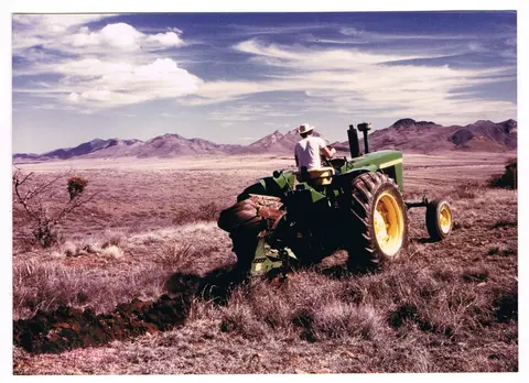 A farmer driving a tractor plowing a field in an arid landscape with mountains in the background.