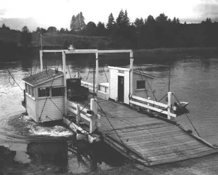 A historic black and white photograph of a small vehicle ferry on a river.