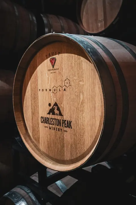 A wooden wine barrel rests indoors in a cellar with other barrels.