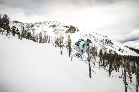 A skier jumps on a snowy mountain slope with trees in the background.